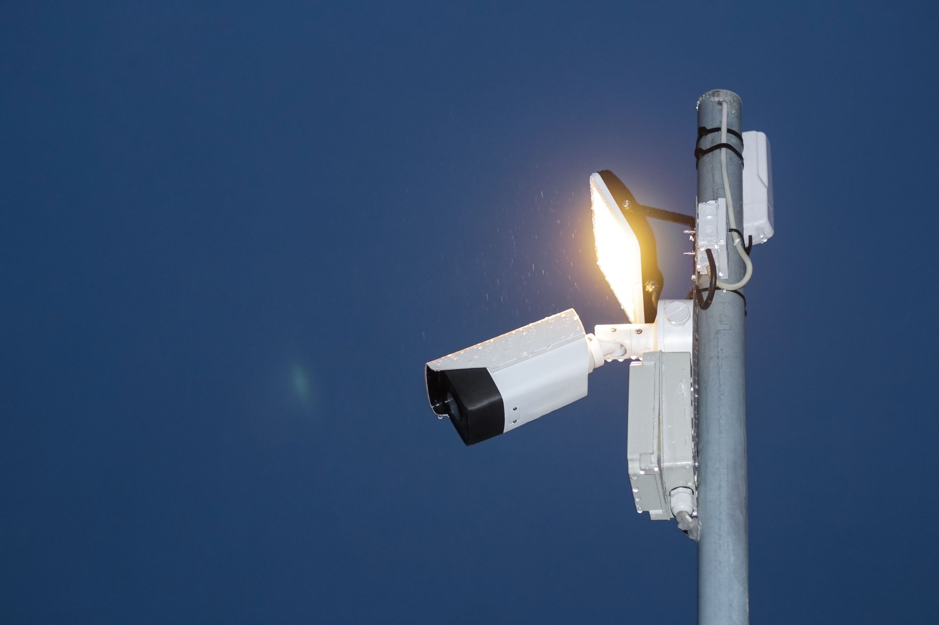 Cámara bullet CCTV exterior en poste con panel de luz blanca encendido contra cielo nocturno azul oscuro