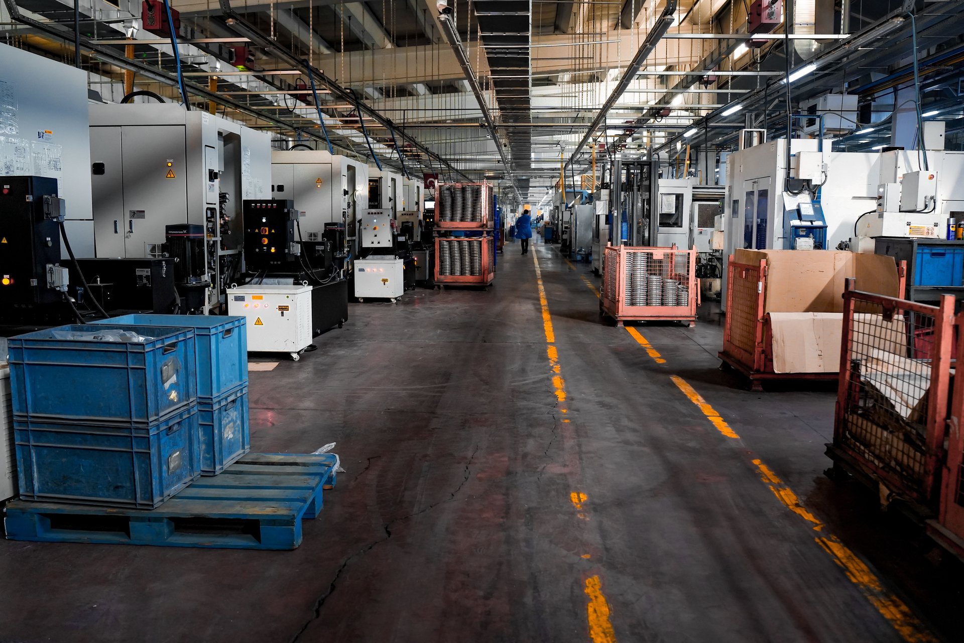 Pista de piso de planta industrial con maquinaria pesada a ambos lados, marcado amarillo de seguridad en el suelo, cajas azules y racks metálicos
