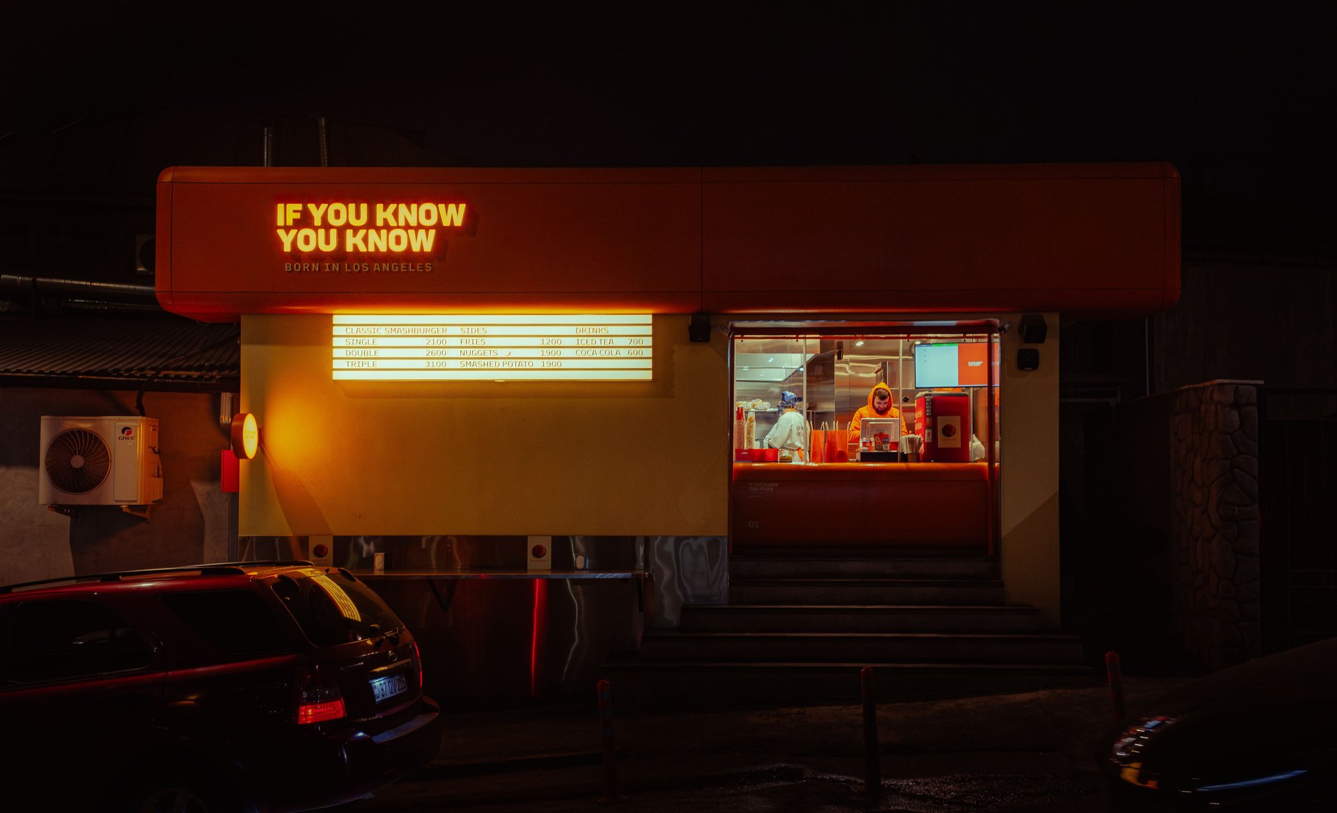 Ventanilla de drive-thru de comida rápida iluminada con neón por la noche, con un trabajador visible dentro y un coche esperando en el carril