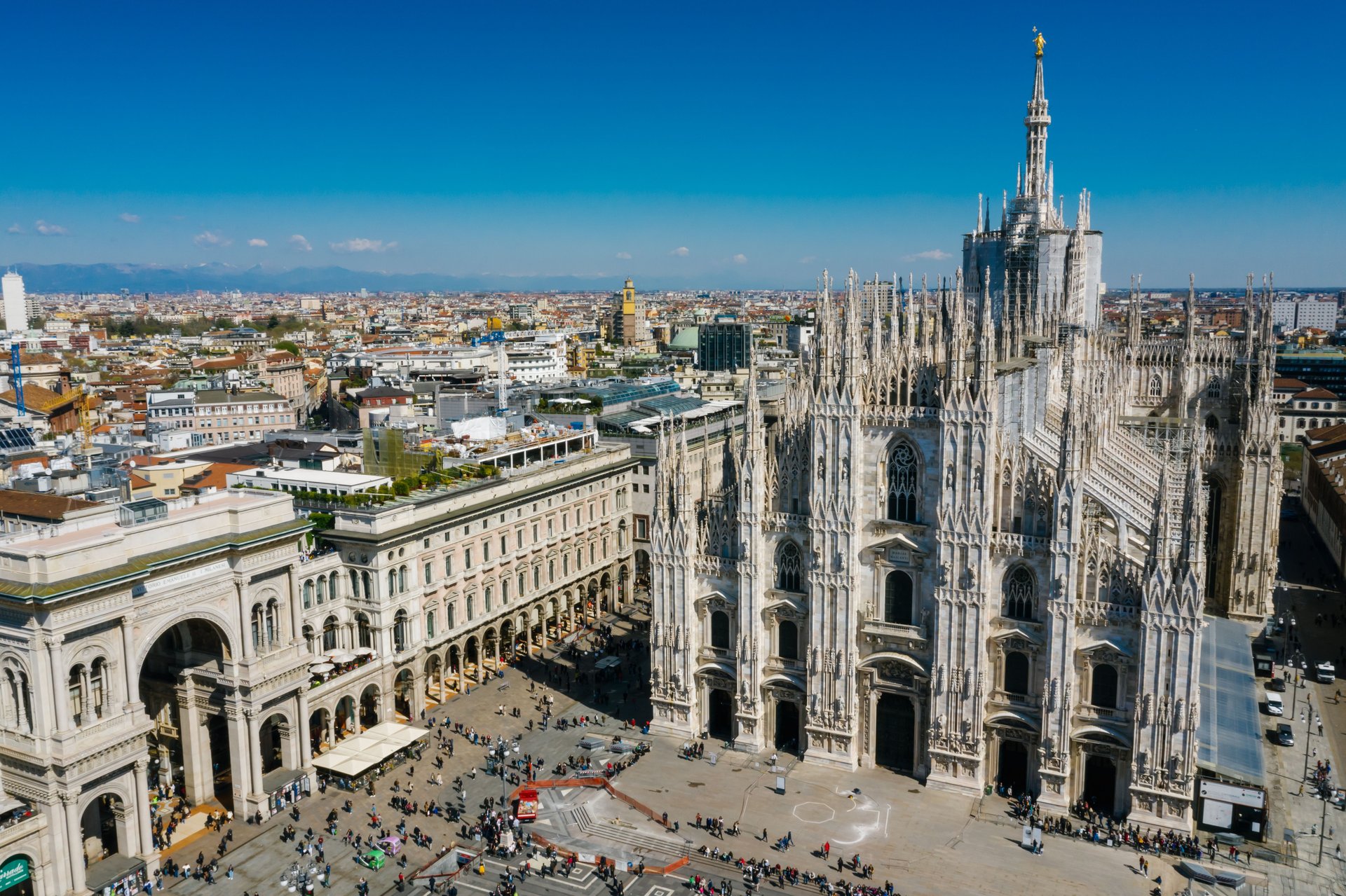 Vista desde el interior de la Catedral del Duomo en Milán a través de la red protectora de la aguja, mirando hacia la Piazza del Duomo con multitudes pequeñas de transeúntes al fondo, sin logos de vendor, sin texto visible, personas no identificables — visualizando el 'sensitive site' y la arquitectura de vigilancia que el Decreto 160 busca regular.