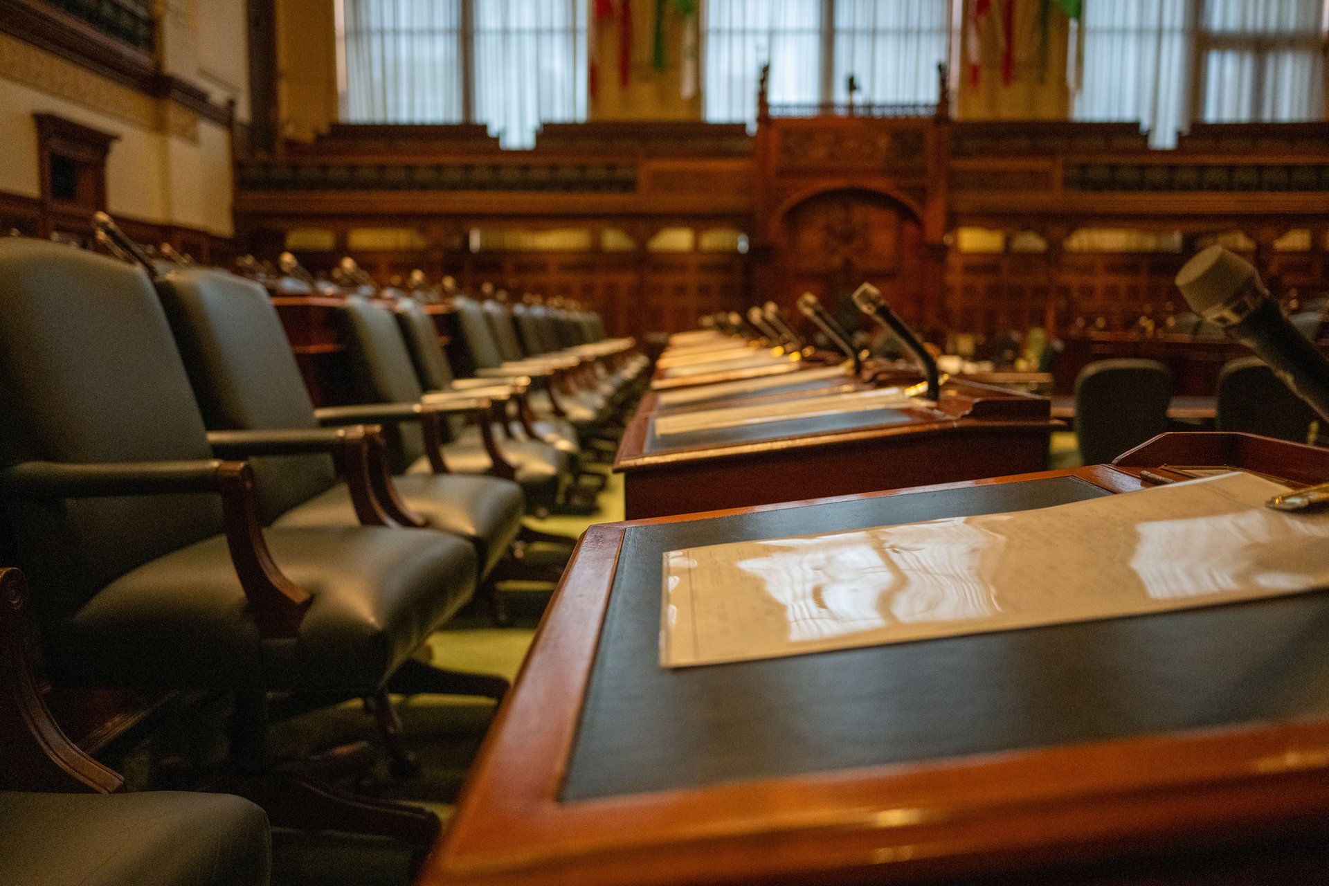 Sala legislativa vacía con filas de escritorios de madera y micrófonos — el espacio donde el voto 5-3 envió la revisión del contrato Flock a comité sin comentario público